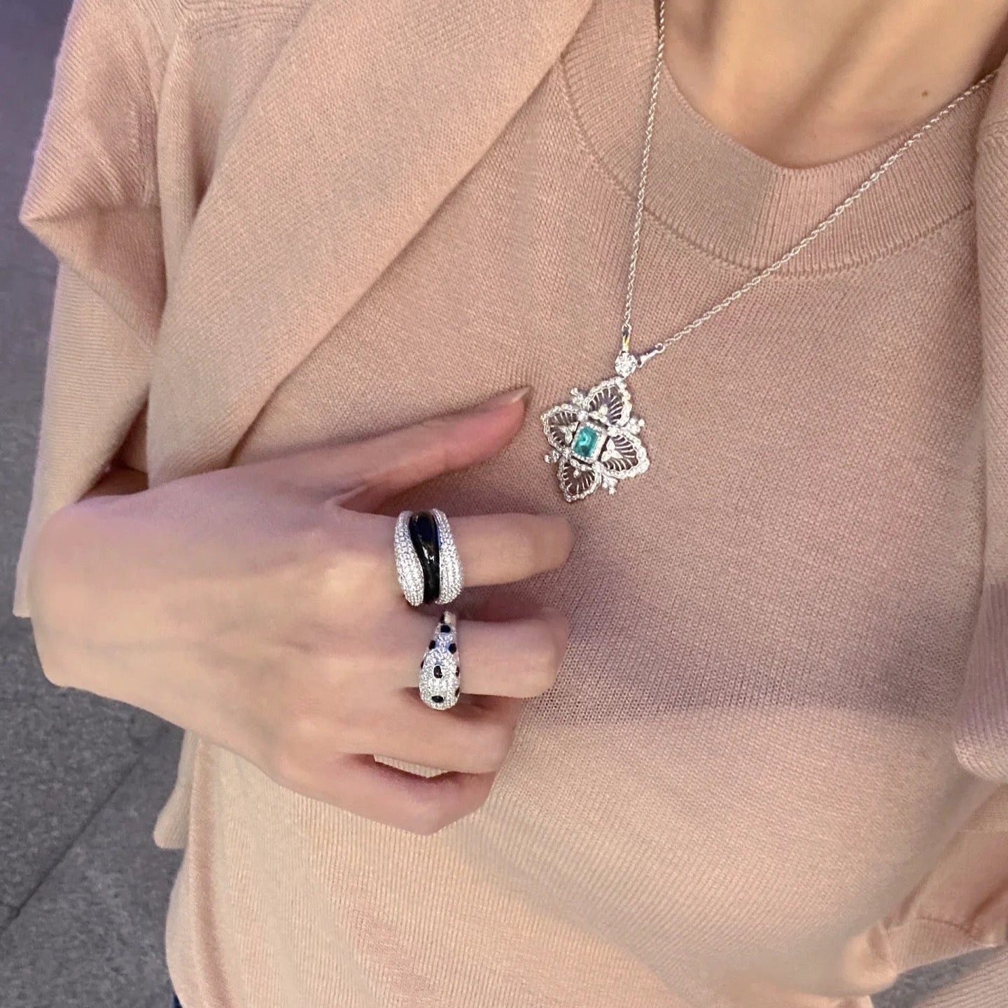 Person wearing a beige jacket with a necklace and rings, standing on a gray pavement.