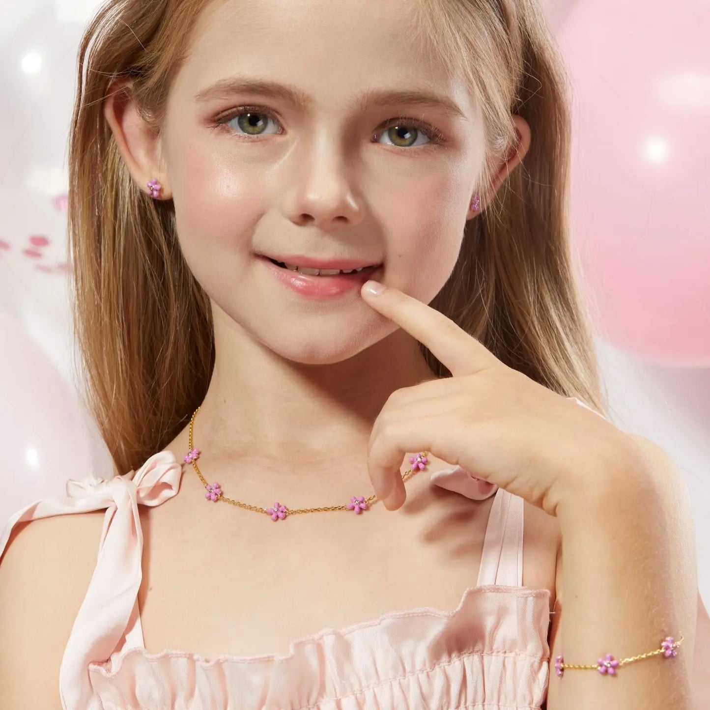 Young girl wearing a pink dress with ribbons and a necklace, surrounded by balloons.